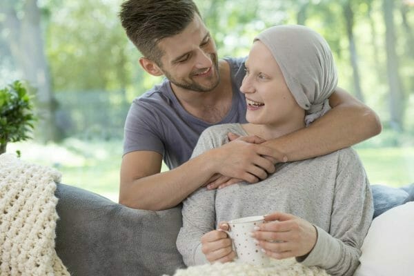 A young woman wearing a headscarf, showing she is undergoing cancer treatment, sits on a couch holding a mug and smiling. A man, likely her partner, hugs her from behind, offering support. They are in a bright, cozy living room with a view of green trees outside.