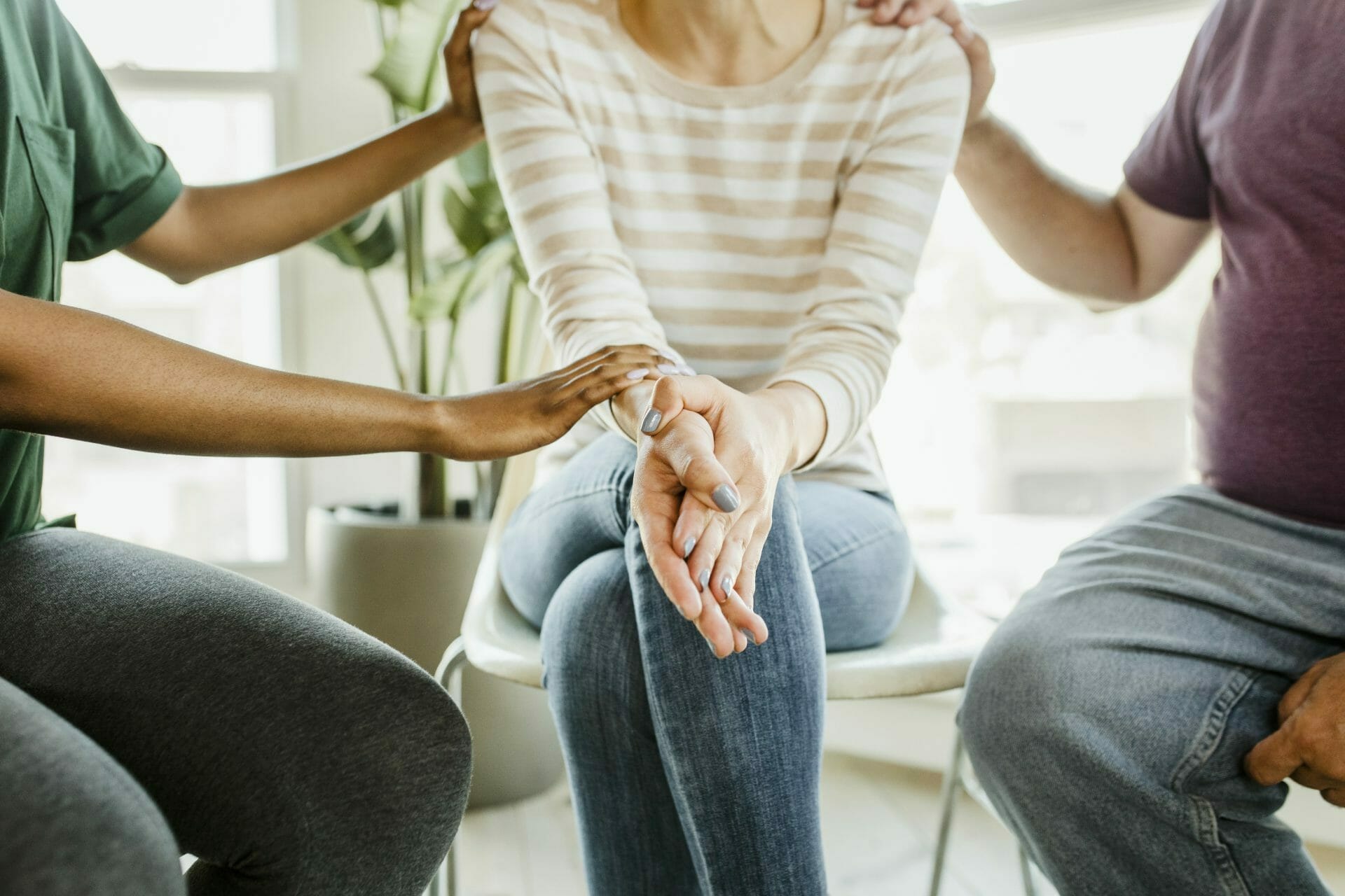 A person in a support group is comforted by others touching their arms and shoulders. The setting is warm and inviting with natural light and a plant.