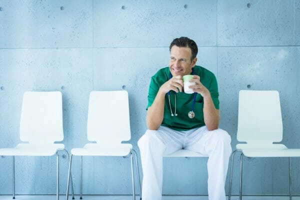 A doctor wearing green scrubs and a stethoscope around his neck is sitting on a chair in a waiting area. He is smiling and holding a cup of coffee, taking a break. The background is a light blue wall with a modern, minimalist design.