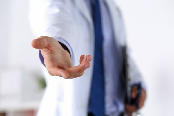 A doctor wearing a white coat and a blue tie extends a hand forward, offering help or assistance. The background is out of focus, emphasizing the gesture of support and care.