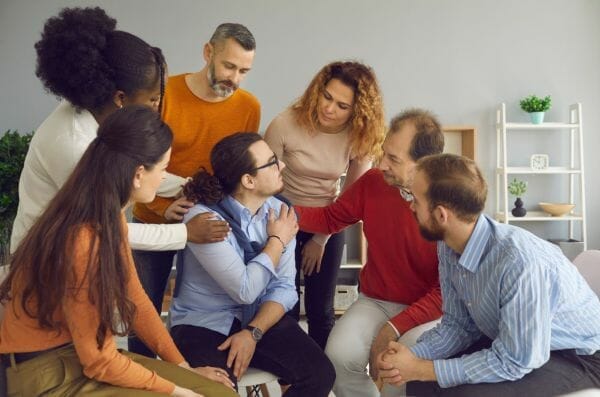 A support group gathers around a distressed man, offering comfort by touching his shoulders. The setting is a warm, welcoming room, symbolizing support for cancer-related financial issues.