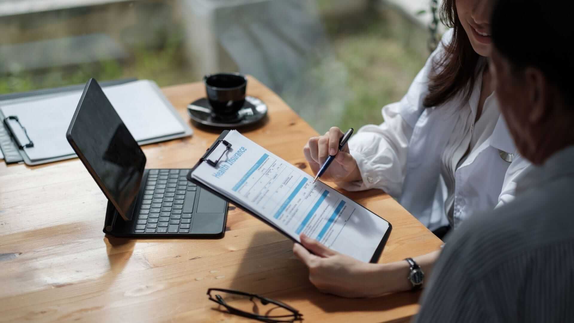 Two individuals in a meeting, with one reviewing a health insurance form that could assist with covering skin cancer treatment expenses. The setting includes a wooden desk with a laptop, clipboard, and other work-related items, suggesting a professional consultation.