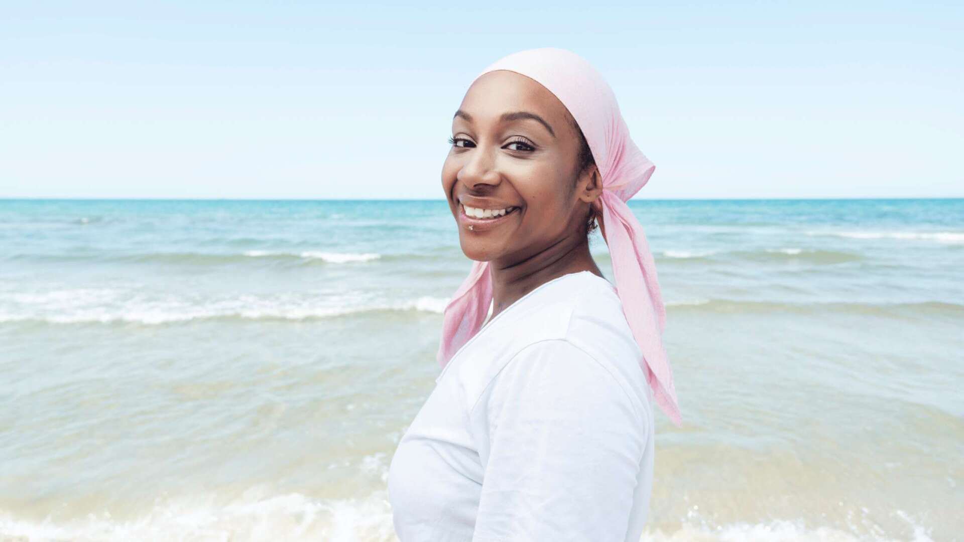 Happy woman in a pink headscarf standing by the beach, representing peace and relief through viatical settlements.